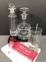 Full set of two glass decanters, martini pitcher, glass stopper, and wine towel displayed on black surface with gray background to show all items.