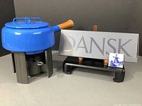 Full lot with blue enamel Dansk fondue pot on black cast iron base, including attached fuel cell, set of 6 fondue forks in box, sign with Dansk branding, and two small Delft tiles with windmill scenes displayed on a stand.