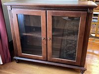 Front view of wooden glass front cabinet showing double glass doors and interior shelves on hardwood floor.