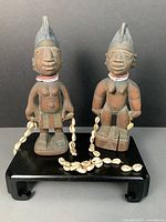 Front view of both carved Ibeji figures on black stand showing beaded collars and shell garlands