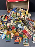 Wide view of over 50 vintage paper match books displayed loosely with a cardboard box in the background.