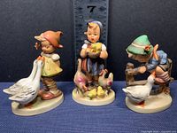 Front view of three M.J. Hummel figurines including Goose Girl, Feeding Time, and Barnyard Hero showing details and size scale
