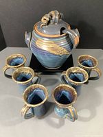 Full view of tureen with lid and ladle, surrounded by six mugs from above showing interior and blue swirl glaze.