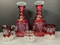 Photo showing full set of two cranberry glass decanters with etched grape design, small cup, creamer, and sugar bowl, all against dark background.