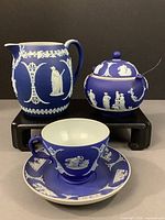 Photo showing three-piece Wedgwood Jasperware tea set including creamer, lidded sugar bowl with spoon, and teacup with saucer, all cobalt blue with white classical figures and patterns.
