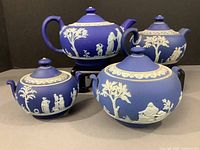 Four blue Wedgwood jasperware pieces including two teapots and two sugar bowls, each decorated with white classical figures and leaf pattern around the rims.