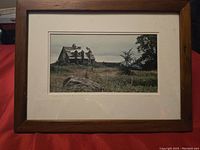 Front view of framed photo showing a rustic house in a field under cloudy sky, framed in wood with beige mat.