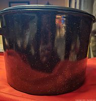 Close-up photo of black enamel-speckled metal pot with matching lid on red surface