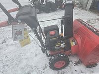 Front-angle view of snow blower on snow-covered ground, showing housing, engine, and control handles.