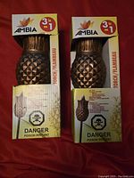 Photo showing two boxed Ambia Tahiki pineapple-shaped torches in original packaging with warning labels visible.