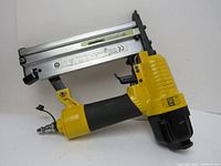 Right-side view of yellow and black air stapler showing the trigger, grip, metallic staple magazine, and air hose connector.