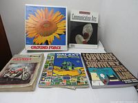Books arranged on flat surface showing covers of Ground Force, Communication Arts, Honda motorbike repair, Galaxy World stamp book, and comic book.