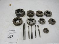 Photo showing 10 large threading dies and 4 screw taps for threading operations, metal with surface rust.
