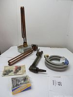 Two copper heating elements, a dishwasher connection hose, a valve, a torch, and packets of hardware arranged on a table with white backdrop.