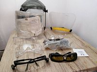 Set of safety eye protection gear including two full face shields, several pairs of safety goggles, and LED inspector glasses on a wood surface.
