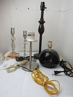 Lot of five table lamp bases arranged on white surface. Two vintage bases: one glass crystal with metallic socket, one dark hard plastic rounded base with gold socket. Three newer bases include brushed metal cylindrical base, two clear glass bases—one segmented and one rounded with wooden base. All without lampshades and with cords attached.