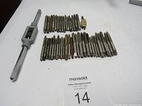 Photo showing 46 metal taps laid out in groups with a single metal tap handle on a white surface. Some taps show signs of surface rust.