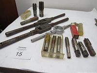 Overall view of 9 large taps, 3 tap handles, and 1 milling machine bit arranged on a white surface. Mixed used and new condition, some showing surface rust.