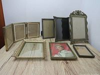 Eight assorted metal picture frames displayed on light wood surface showing various designs including three-fold frame, and frames with floral and striped decorative accents.