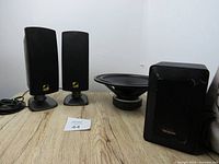 Photo showing GENEXXA bookshelf speaker pair on stands, Peerless 10 inch woofer, and Cyber Acoustics speaker with metal grille on a wooden surface.