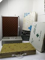 Seven vintage metal and plastic boxes consisting of two first aid kits, one light briefcase, two metal money boxes, one rusty green metal box and one plastic game box, all showing varying levels of wear and rust.