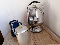 Photo of three coffee percolators on a wooden surface against a white wall showing the large Samovar style percolator, silver camping percolator, and blue camping percolator