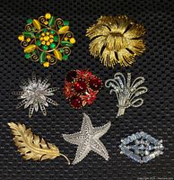 All seven vintage brooches displayed on black textured background showing a variety of shapes, colors and designs.