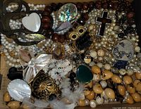 Top view of assorted costume jewelry in a box showing multiple beaded necklaces, pendants including a black cross, an owl brooch, and mother-of-pearl flower brooch.