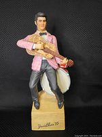 Front view of ceramic Elvis Presley liquor decanter in pink jacket holding guitar with drum set in the background.