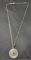 Full view of the sterling silver box chain necklace with attached circular pendant resembling a vinyl record, lying flat on a dark fabric background.