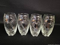 Four clear textured glass drinking glasses embossed with cat faces on front and tails on back arranged side by side against a black textured background.