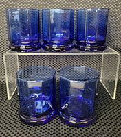 Five cobalt blue old fashioned glasses with 10 sides and thick bases, arranged on display stand.
