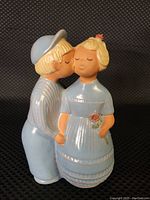Full side view of the light blue ceramic figurine showing the boy kissing the girl on the cheek, both in traditional attire. Figurine measures approximately 7" tall.