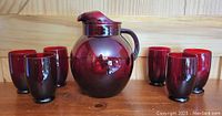 Group photo showing the ruby red glass water jug with six matching glasses arranged around it on a wooden surface with a wood panel background.