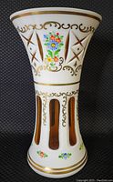 Full side view of the vase showing the milk glass body with multiple cutouts revealing amber glass underneath, decorated with a painted gold scroll pattern and colorful floral painting.