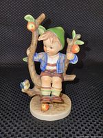 Front view of the Hummel figurine showing the boy sitting in the apple tree holding branches with apples and a small bird on the tree trunk.
