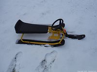 GT Snower Racer snow sled standing on snow with yellow frame and black padded seat.
