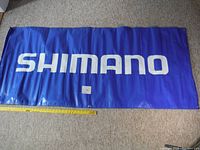 Full view of the blue vinyl banner with white SHIMANO text displayed flat on the floor with a measuring tape showing size.