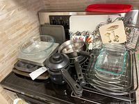 Photo showing multiple kitchen bakeware and cookware items including Pyrex and unbranded glass baking dishes, metal muffin trays, saucepans and trivets arranged on a stove.