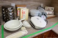 Overview of baking and cooking supplies arranged on a kitchen counter showing all items in the lot including the spice rack, colanders, plastic spoon rests, cookbooks, George Foreman grill and kitchen linens.