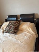 Front view of queen size bed with dark brown leather headboard and storage drawers at foot, covered with light colored bedding and a decorative pillow.