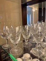 Multiple clear cut glasses with etched cross and olive pattern in a display cabinet, varying sizes including wine glasses and brandy snifters. Some glasses have taped markings indicating chips.