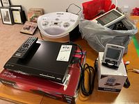 Sony DVD player DVP-SR510H on its box with remote on top shown with RCA portable CD player and digital travel clock on the side