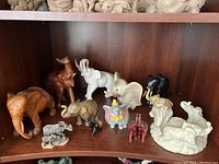 Shelf displaying a dozen elephant figurines of different sizes and materials including wood, ceramic, resin, and stone.