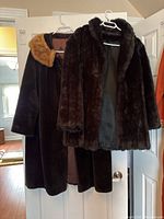 Two coats hanging on hangers on a door: one full-length dark brown coat with tan fur collar, one shorter dark brown faux fur jacket.