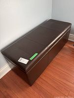 Main photo of brown faux leather storage bench showing top and side details, set on wood flooring near wall.