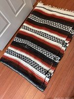 Full view of the folded handmade blanket on wooden floor showing the striped and geometric diamond pattern in rust, black, and cream colors.
