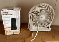 Photo of the white and black electric space heater in original box beside the white Hawaiian Breeze fan on a wooden surface.