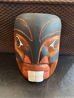 Front view of hand carved and painted wooden mask with black, orange, and white design elements, showing stylized facial features and visible wood grain.