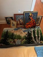 Photo of large hooked rug with nature scene and five framed cross stitch pieces leaning against a wall